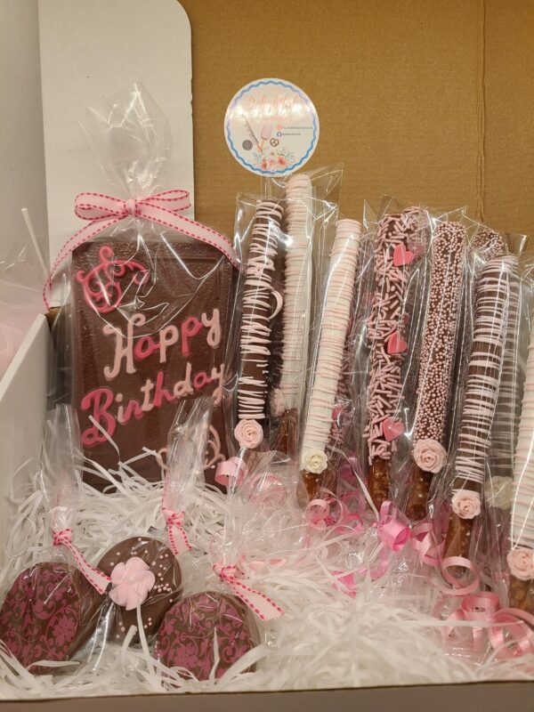 The Large Chocolate Treats Happy Birthday Box includes a "Happy Birthday" chocolate plaque, decorated pretzel sticks, and chocolate-dipped items wrapped with pink bows and accents.