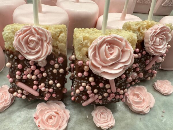 Chocolate-covered Rice Krispie treats adorned with pink candy roses and sprinkles, surrounded by pink cake pops in the background. Perfect for shower, wedding, or birthday party favors.