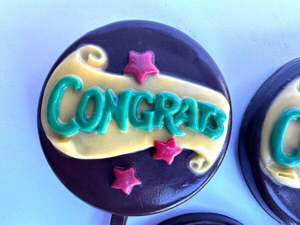 A "Congratulations" chocolate lollipop featuring green icing on a yellow banner with red stars against a dark backdrop.