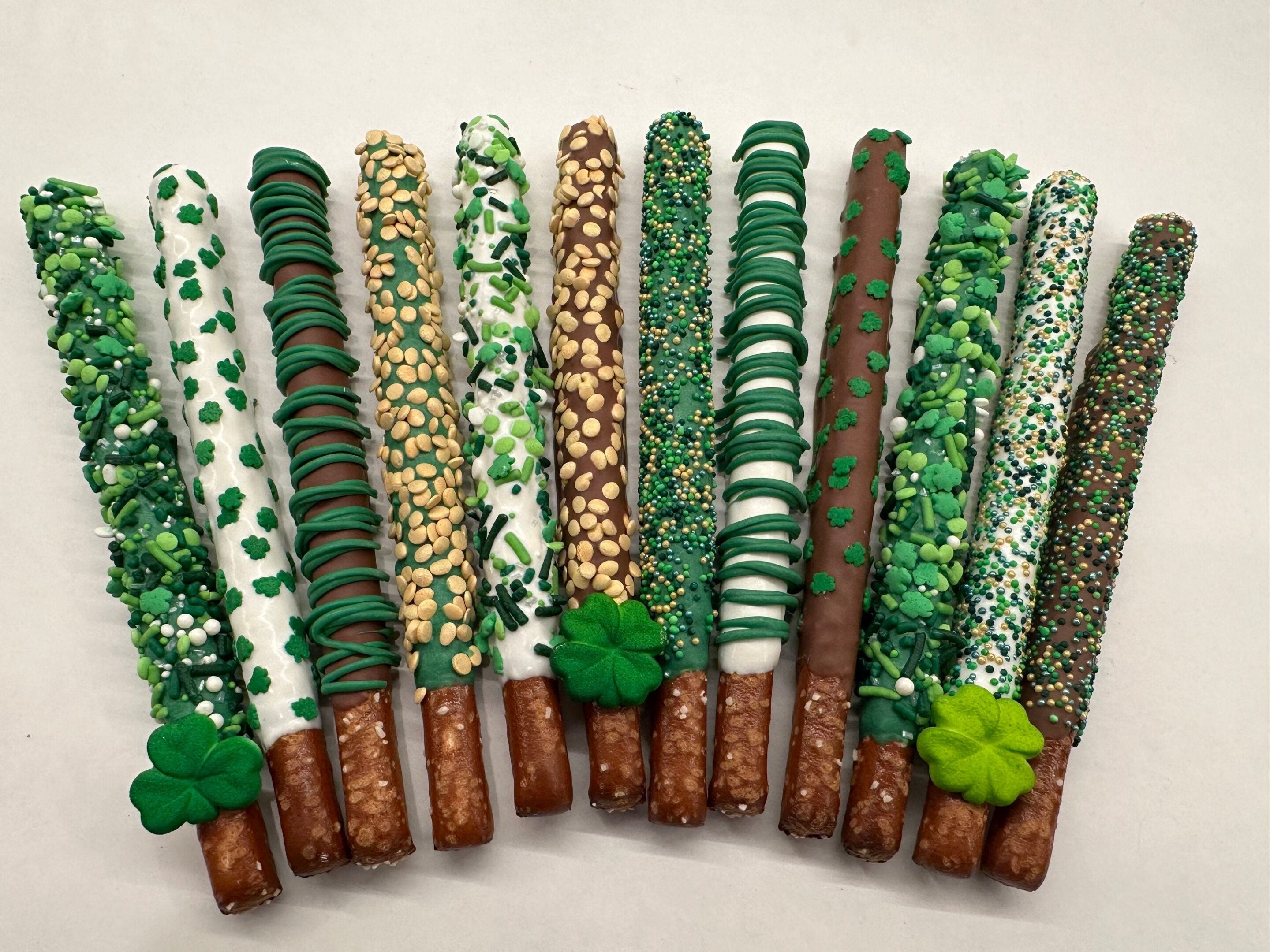 Twelve St Patrick’s Day Pretzels, chocolate-coated and adorned with green and white sprinkles, nuts, and shamrock shapes, displayed on a white background.