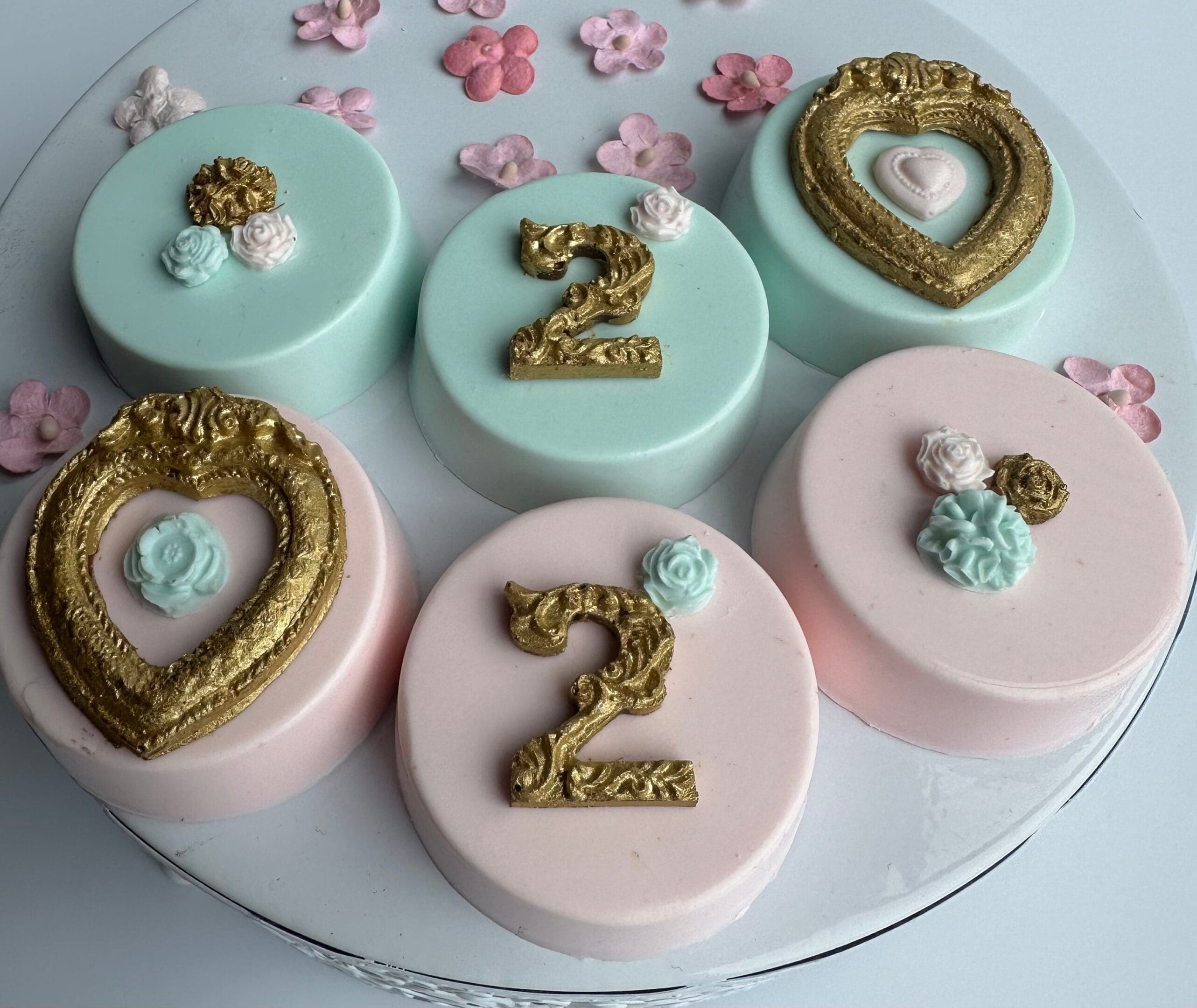 Six decorated cakes featuring pastel colors, heart-shaped ornaments, and "2" toppers, alongside 12 Gold Frame Chocolate Covered Oreos for a girl's or boy's birthday with age, all surrounded by small floral decorations on a white surface.