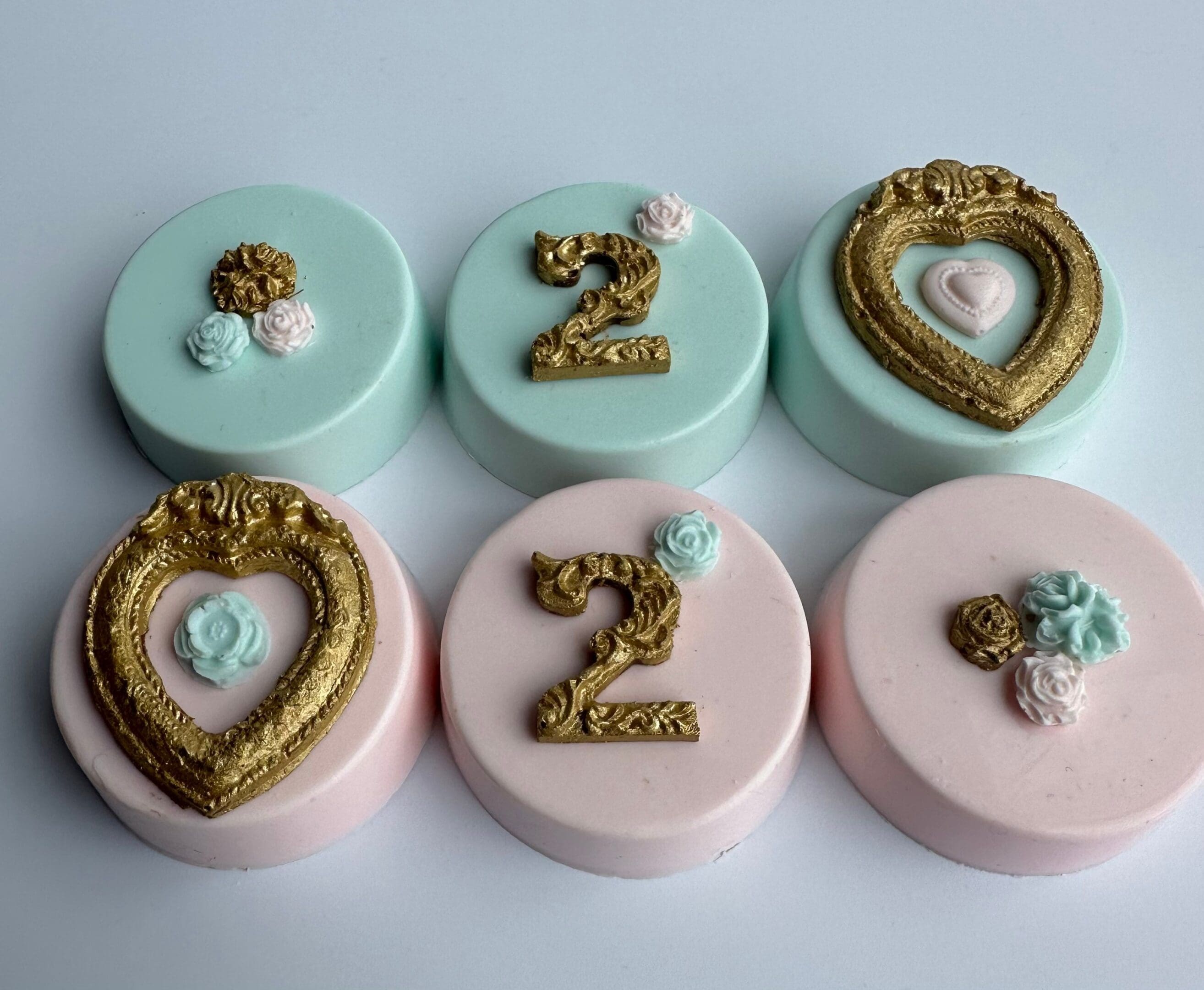 Five chocolate-covered Oreos featuring roses, two in light green and three in pink, each adorned with a gold heart frame or the number "2".