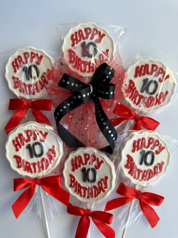 A set of six white chocolate lollipops featuring red "Happy 10th Birthday" text and decorated with red bows is arranged around a black and red ribbon centerpiece. Product: 12-Happy Birthday Chocolate Lollipops With Age.