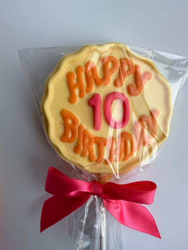 A round cookie in the shape of a lollipop, inscribed with "Happy 10 Birthday," wrapped in plastic, and tied with a pink ribbon is part of the 12-Happy Birthday Chocolate Lollipops With Age set.