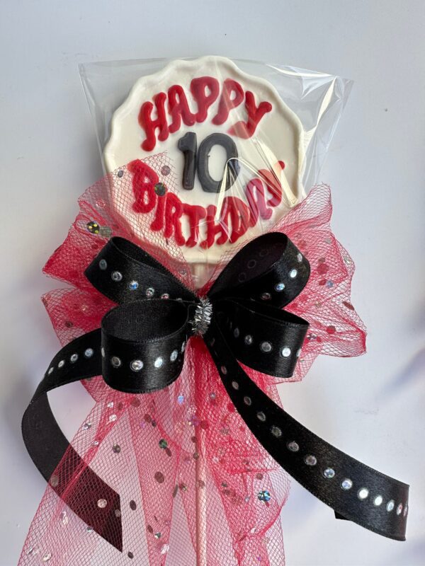 A "12-Happy Birthday Chocolate Lollipop With Age," featuring "Happy 10 Birthday" in red and black icing, wrapped in plastic, and decorated with a red netting and black ribbon with silver studs.