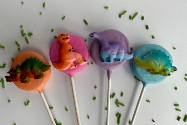 A set of 12 Dinosaur Chocolate Covered Oreo Pops, featuring colorful dinosaur designs on a white background, complemented by scattered green sprinkles.
