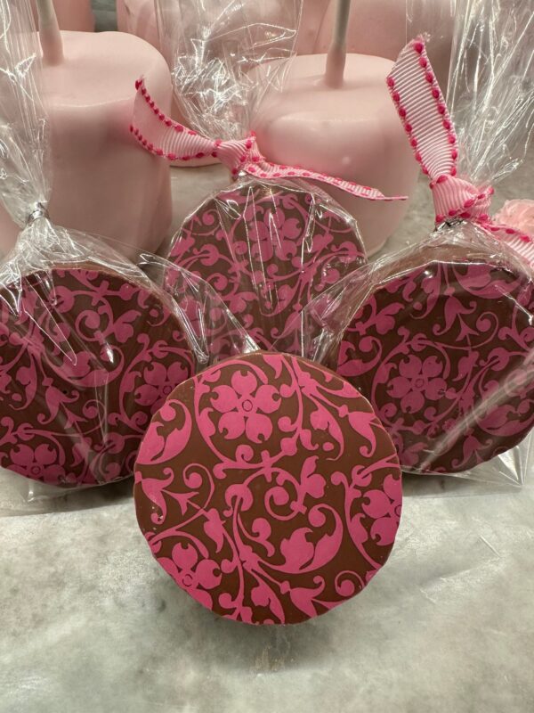 The "12-Pink Floral Scroll Chocolate Covered Oreo Gift Box" features treats adorned with a pink pattern, wrapped in clear plastic and tied with a pink ribbon on a marble surface. Perfect for weddings, birthdays, party favors, or baby showers.