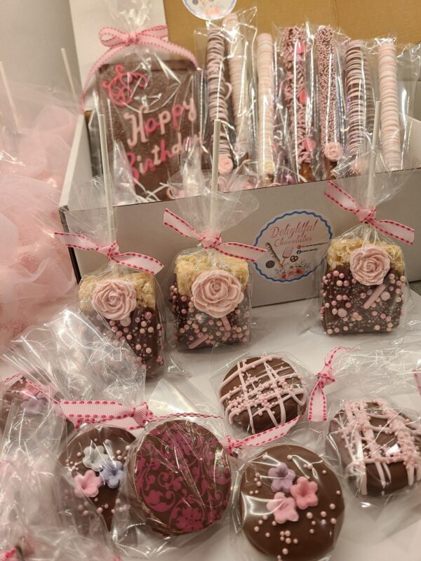 The Large Chocolate Treats Happy Birthday Box includes assorted decorated chocolate treats like cakesicles, cookies, and rice crispy treats in clear packaging with pink ribbons, featuring floral designs and "Happy Birthday" text.