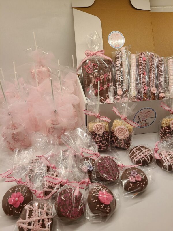A selection of decorated chocolate treats, such as cake pops and pretzel rods, wrapped in pink and clear packaging, is displayed on a surface with the Large Chocolate Treats Happy Birthday Box in the background.
