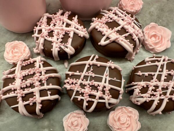 A Large Chocolate Treats Happy Birthday Box features chocolate cookies with pink drizzle and sugar pearls, accompanied by pink meringue roses on a gray surface.