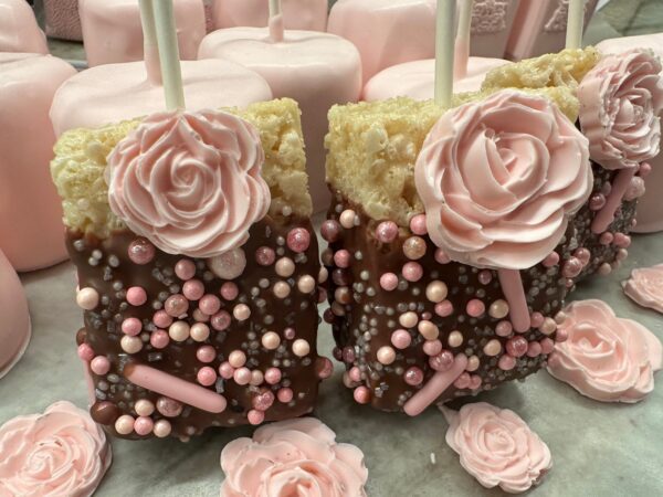 Chocolate-covered Rice Krispie treats with roses, decorated for parties and arranged on sticks, surrounded by pink cake pops.