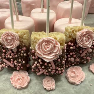 12 Chocolate Covered Rice Krispie Treats with decorative sprinkles and frosting roses are set on a table, accompanied by large pink marshmallows, perfect for shower, wedding, or birthday party favors.