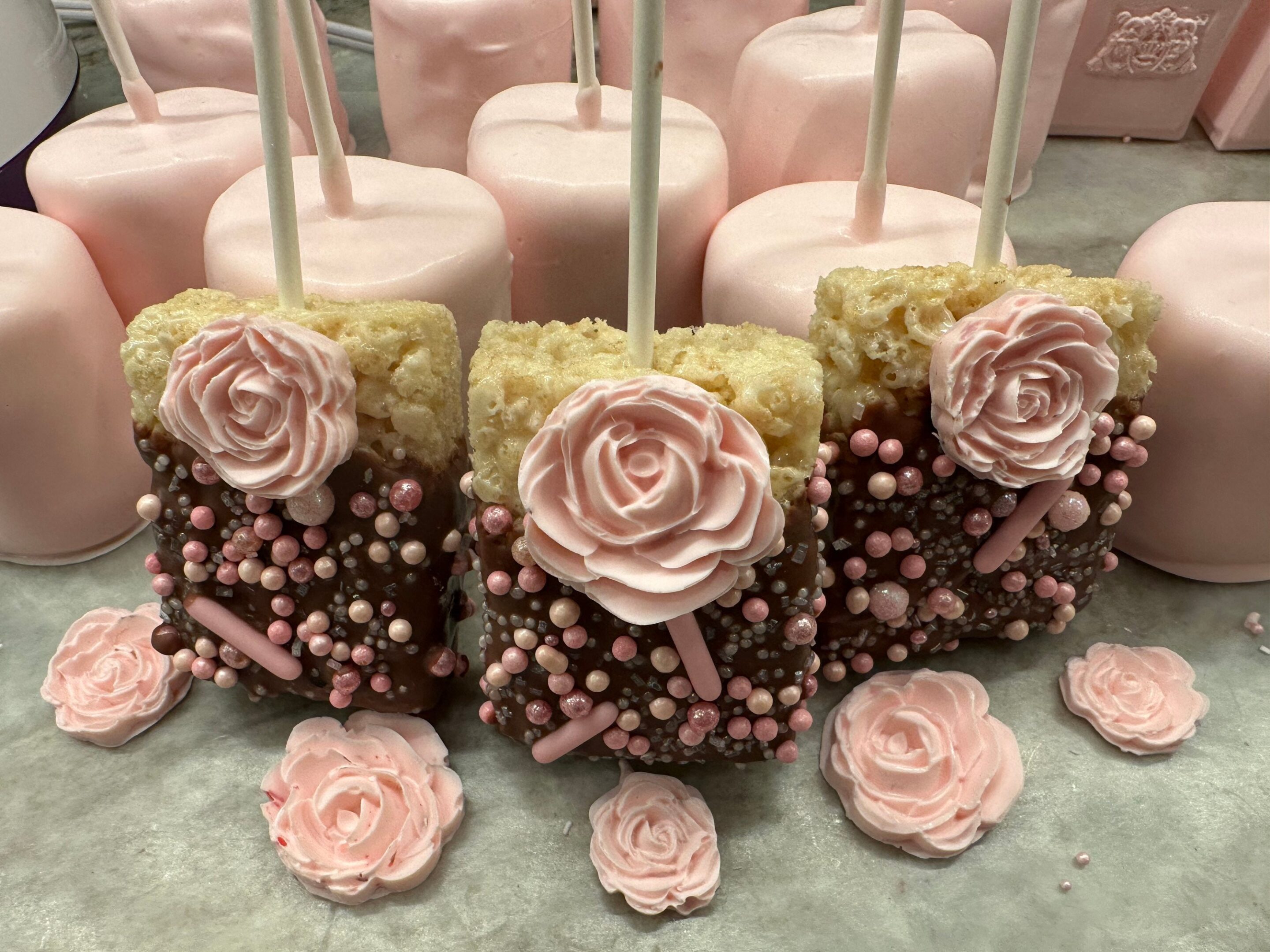 12 Chocolate Covered Rice Krispie Treats with decorative sprinkles and frosting roses are set on a table, accompanied by large pink marshmallows, perfect for shower, wedding, or birthday party favors.