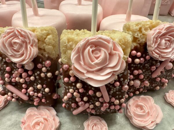 Chocolate-covered Rice Krispies treats with pink icing roses and candy pearls are arranged on a counter as part of the 12-pack party favors for showers, weddings, or birthdays.