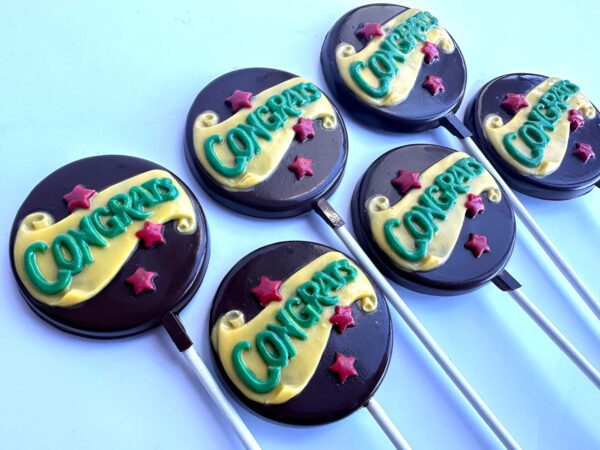 Five chocolate lollipops from the "12-Congratulations Chocolate Lollipops" set feature "Congrats" in green icing with yellow ribbons, adorned with small pink stars on a light blue background.