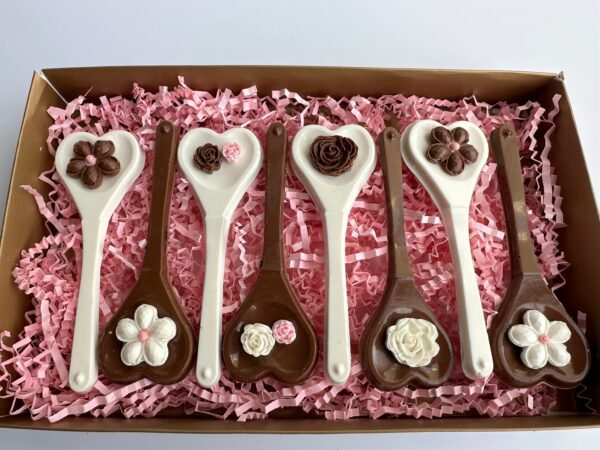 A box of six heart-shaped chocolate spoons with floral designs, arranged on pink shredded paper. Product: Chocolate Heart Spoons Gift Box for Hot Chocolate/Wedding/Birthday Gift/Mother's Day.