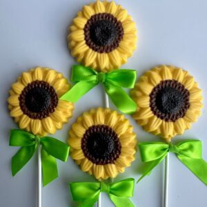 Four sunflower-shaped chocolate lollipops with yellow petals and brown centers, each tied with a green ribbon, on white sticks against a light background.