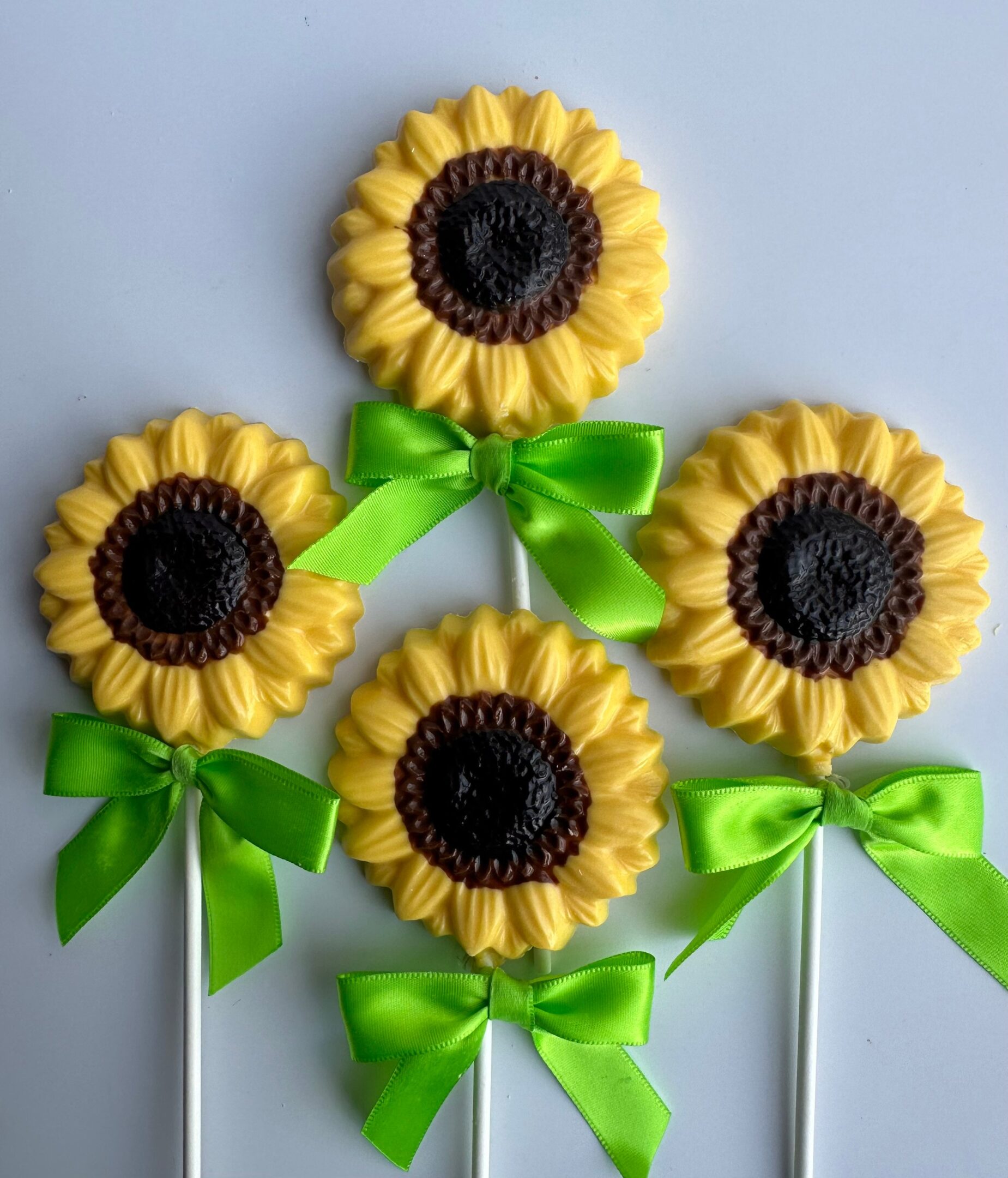 Four sunflower-shaped chocolate lollipops with yellow petals and brown centers, each tied with a green ribbon, on white sticks against a light background.