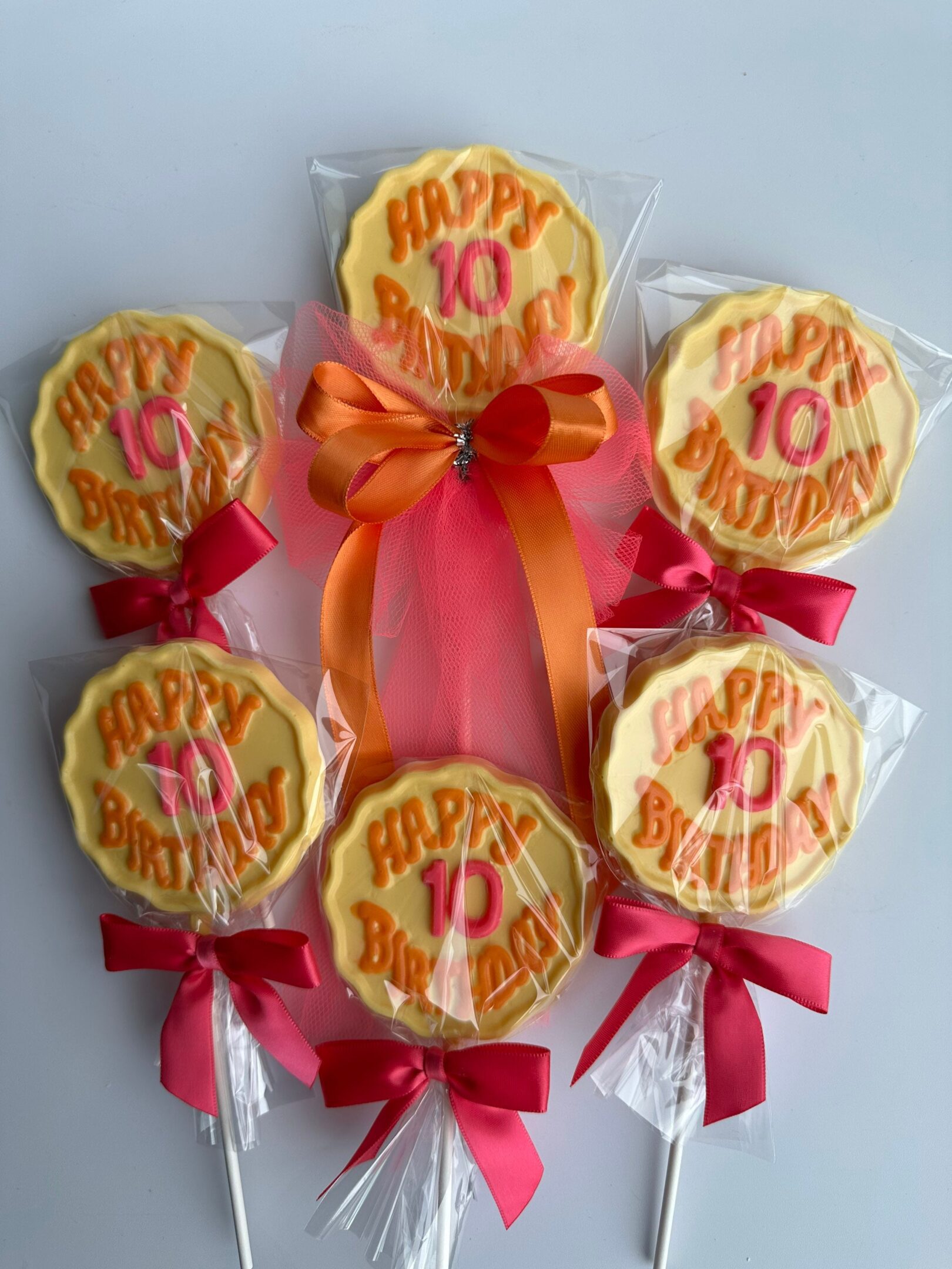 Six "Happy 10 Birthday" chocolate lollipops with yellow cookies, wrapped in plastic with red bows, and arranged in a fan shape around an orange and pink ribbon.
