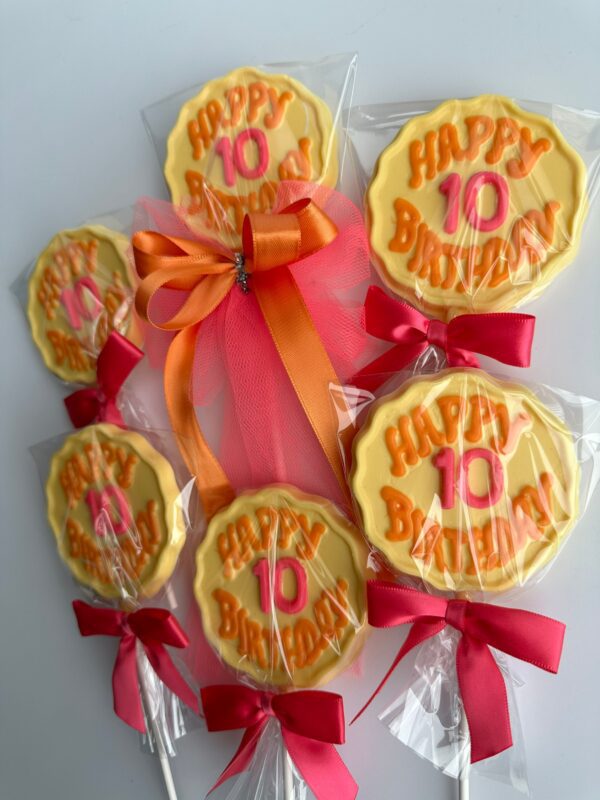 Six "Happy 10th Birthday" chocolate lollipops featuring orange text on yellow discs, wrapped in clear plastic with pink bows at the stick's base, and one adorned with an orange tulle bow.