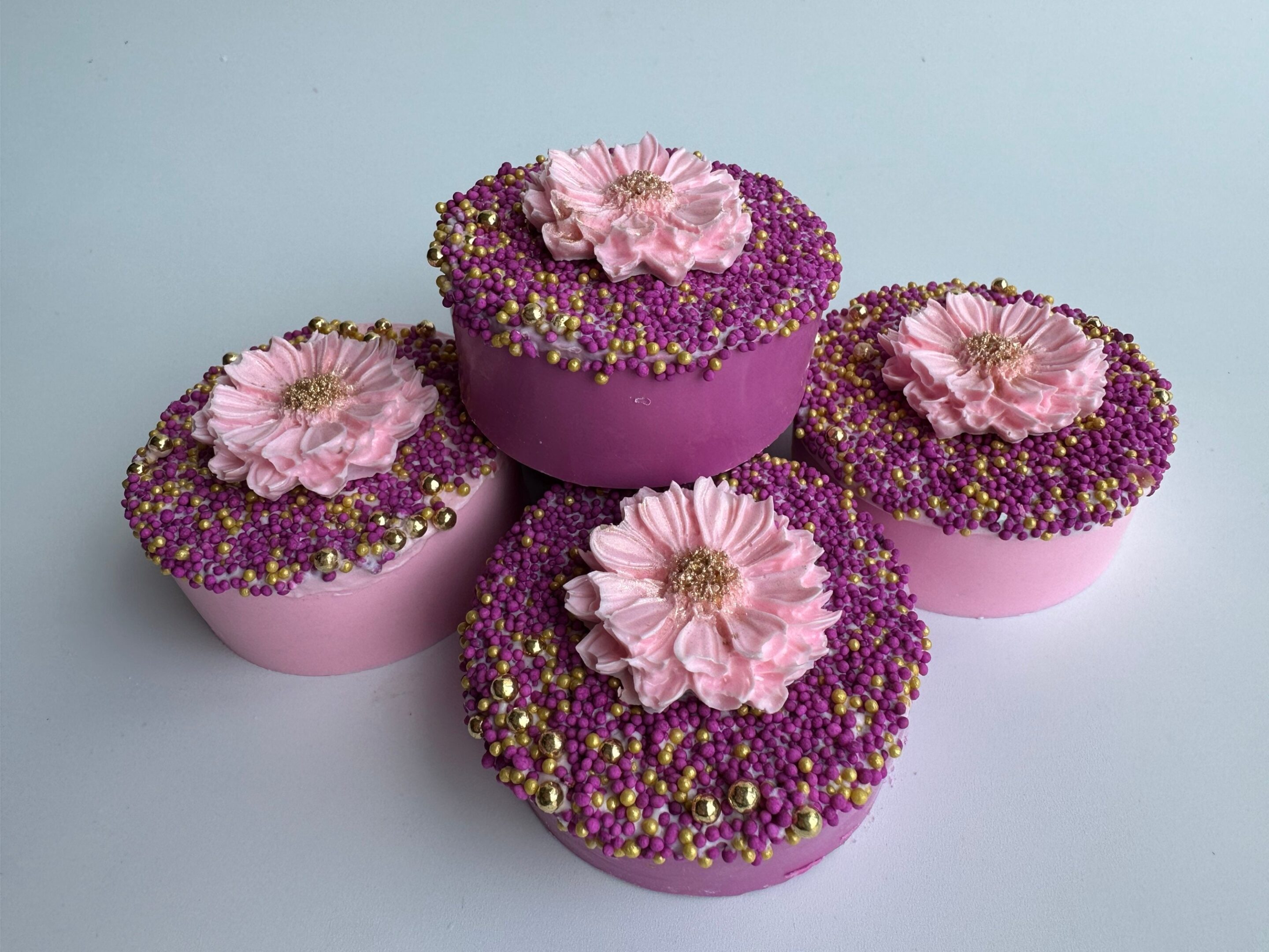 Four decorative cakes with purple and pink frosting, topped with pink flowers and multicolored sprinkles, arranged on a light background.