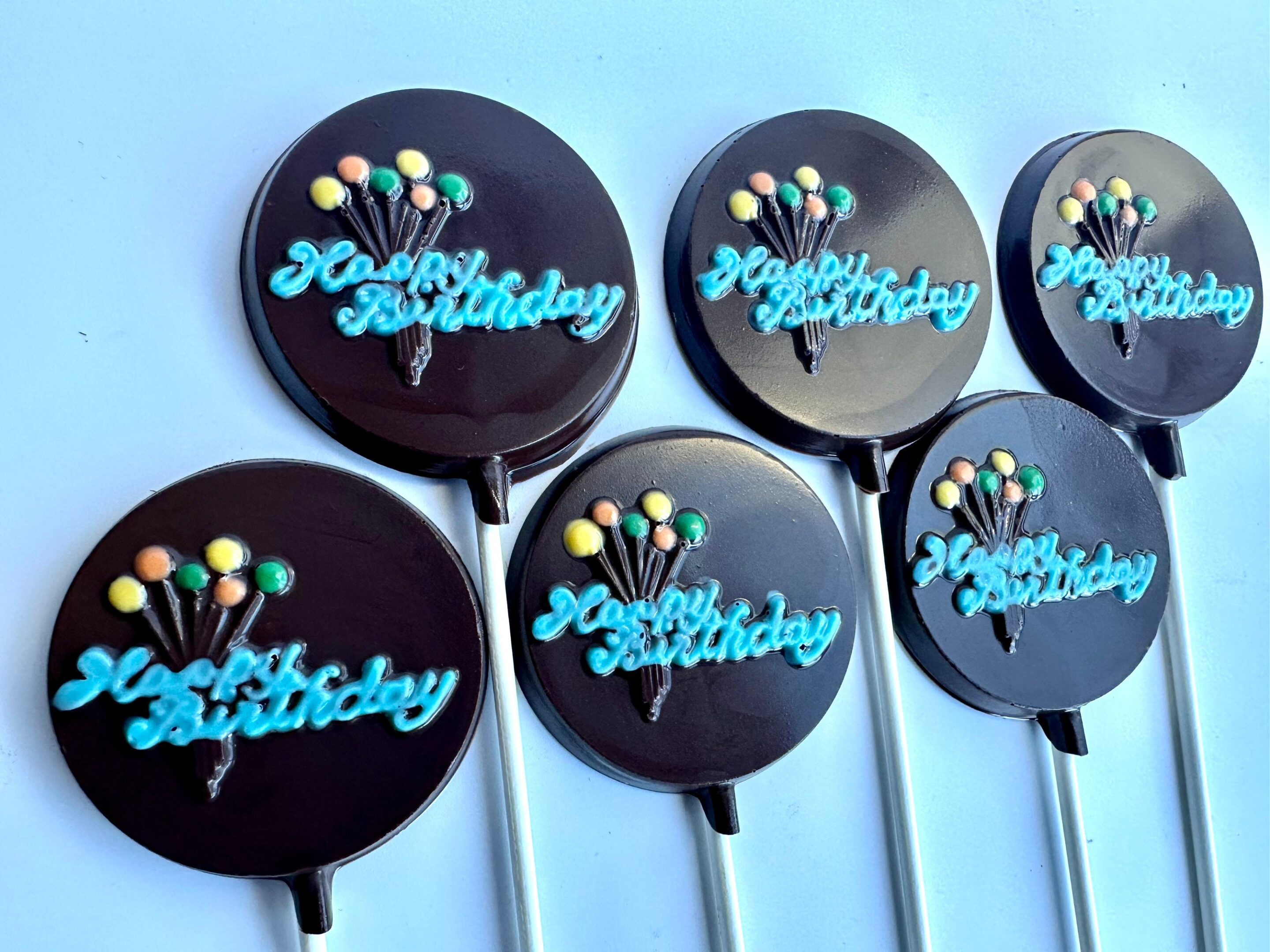 Six "Happy Birthday" chocolate lollipops with blue balloon designs and colorful candy dots.