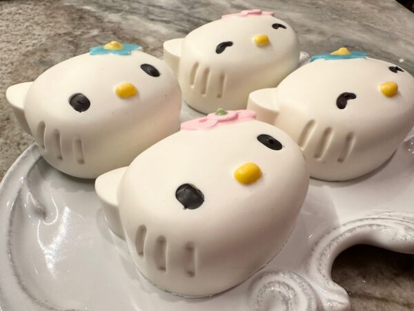 Four chocolate-covered Oreo favors shaped like cartoon cat heads with bows, yellow noses, and black eyes are displayed on a ceramic tray.