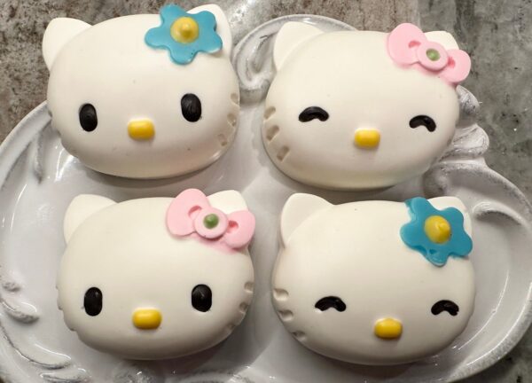 Four chocolate-covered Oreo favors shaped like cats, with white icing and colorful bows, on a ceramic plate.