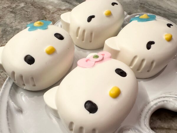 Four whimsical chocolate-covered Oreo kitty favors, with white icing and colorful flower decorations, on a white plate.