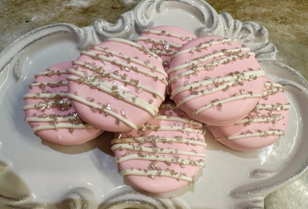 A plate of pastel pink chocolate-covered Oreos with white drizzle, adorned with sugar pearls.