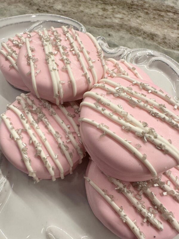 12 pastel pink, heart-shaped, chocolate-covered Oreos with white icing drizzle and silver sprinkles on a decorative white plate.