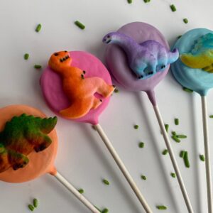 A set of four chocolate-covered Oreo pops shaped like dinosaurs with pink, orange, purple, and blue icing, topped with green sprinkles on a white background.