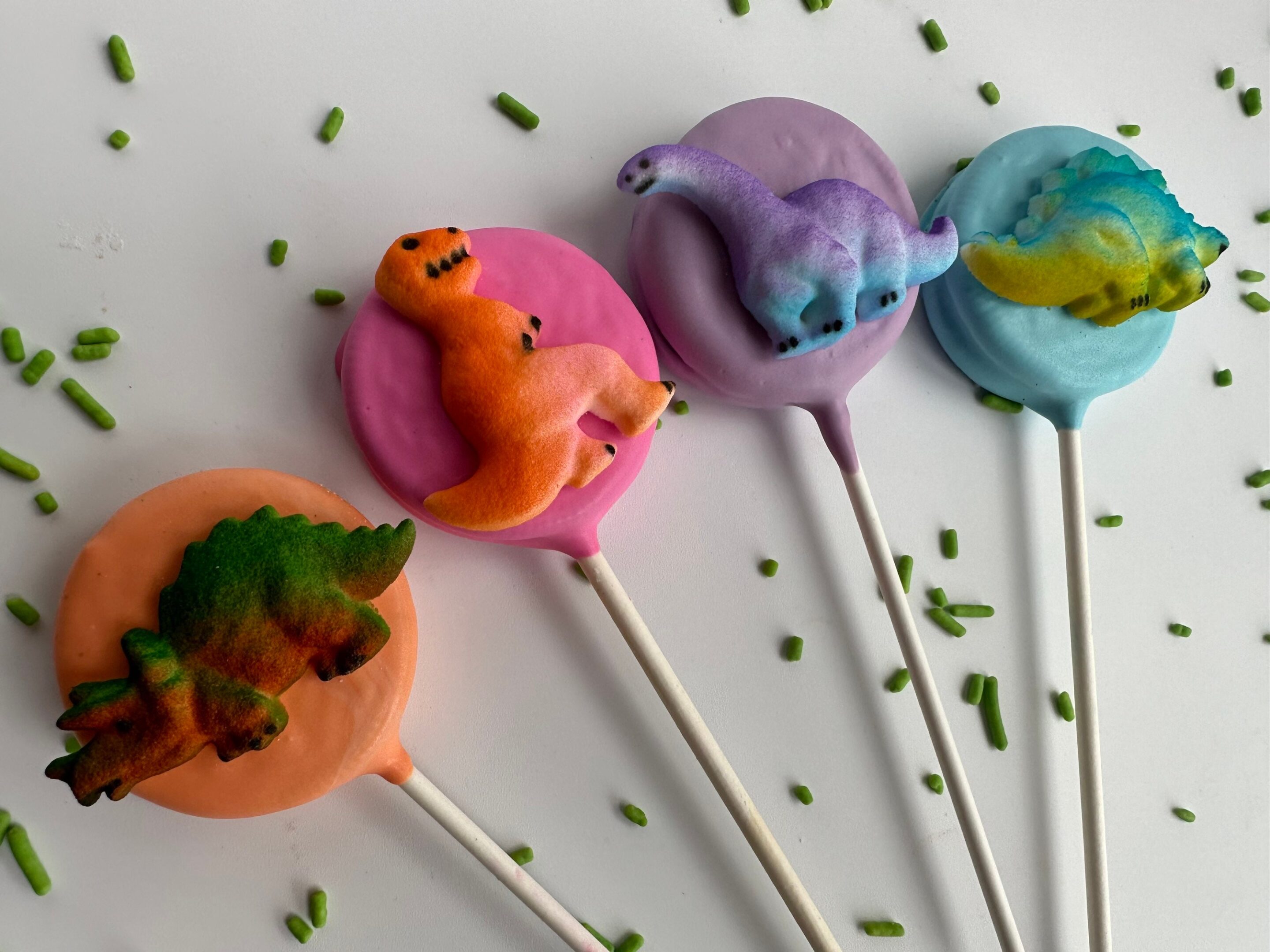 A set of four chocolate-covered Oreo pops shaped like dinosaurs with pink, orange, purple, and blue icing, topped with green sprinkles on a white background.