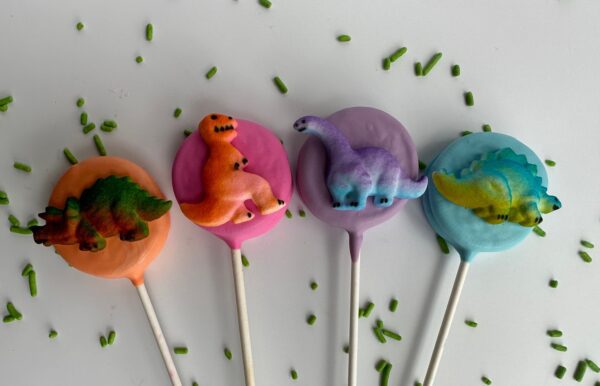 Twelve chocolate-covered Oreo pops topped with small dinosaur figures in orange, pink, purple, and blue, complemented by green sprinkles.
