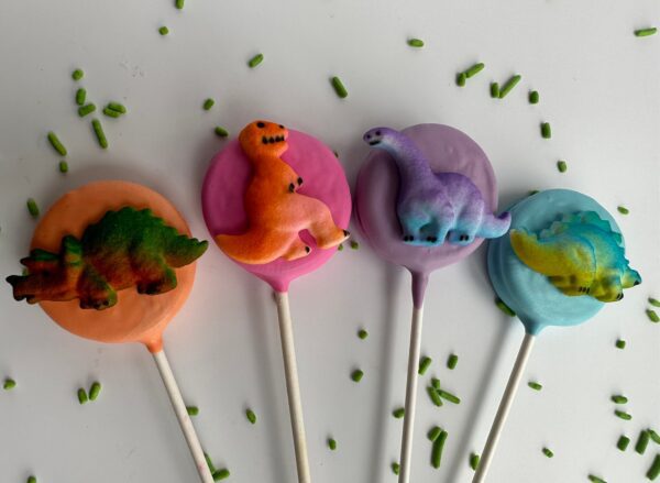 Four vibrant Dinosaur Chocolate Covered Oreo Pops, each featuring a unique dino design, are displayed on white sticks amidst a scattering of green sprinkles.