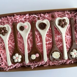 Box containing six chocolate spoons with heart-shaped bowls, decorated with floral chocolate designs, placed on pink shredded paper.