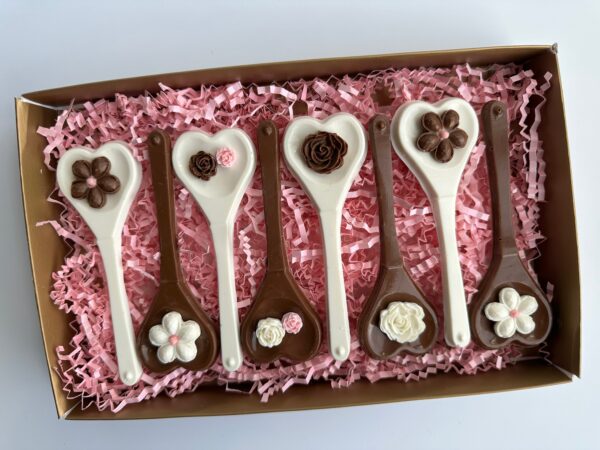 Box containing six chocolate spoons with heart-shaped bowls, decorated with floral chocolate designs, placed on pink shredded paper.