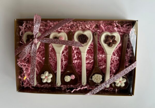 The Chocolate Heart Spoons Gift Box includes six heart-shaped chocolate spoons, each with unique decorations. The box features a lining of pink paper shreds and is tied with a pink glittery ribbon, perfect for hot chocolate or as a gift for weddings, birthdays, or Mother's Day.