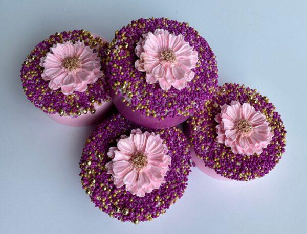 Four chocolate-covered Oreos decorated with pink and purple sprinkles, each topped with a pink icing flower, arranged on a white surface.
