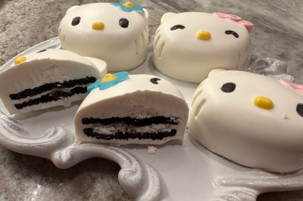 Four chocolate covered Oreo favors, Hello Kitty-themed, are on display: two adorned with blue flowers and two with pink bows. One is sliced to reveal layers of cream and chocolate.