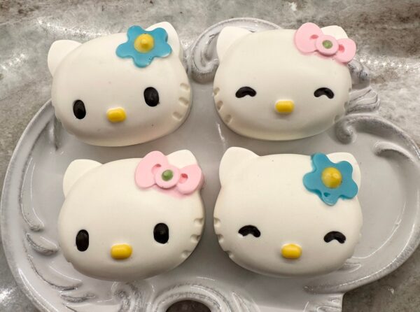 Four 12-cute kitty-themed chocolate-covered Oreo favors with various expressions and decorative bows on a plate.