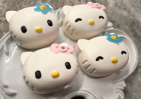 Four chocolate-covered Oreo favors, crafted into white cat shapes with colored bows, elegantly arranged on a decorative white plate.