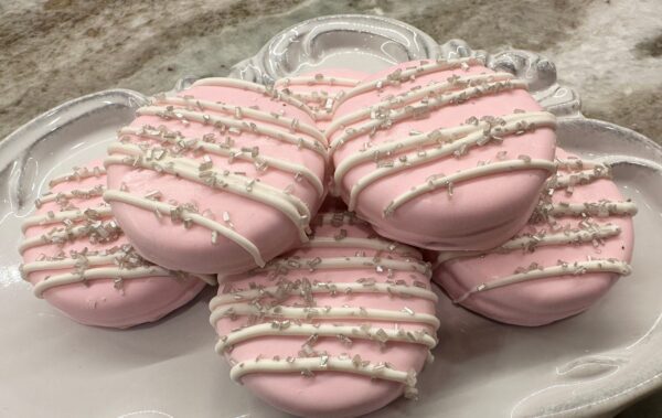Pastel pink chocolate-covered Oreos with white icing and sprinkles on an ornate white plate, available as a 12-pack for weddings or baby showers.