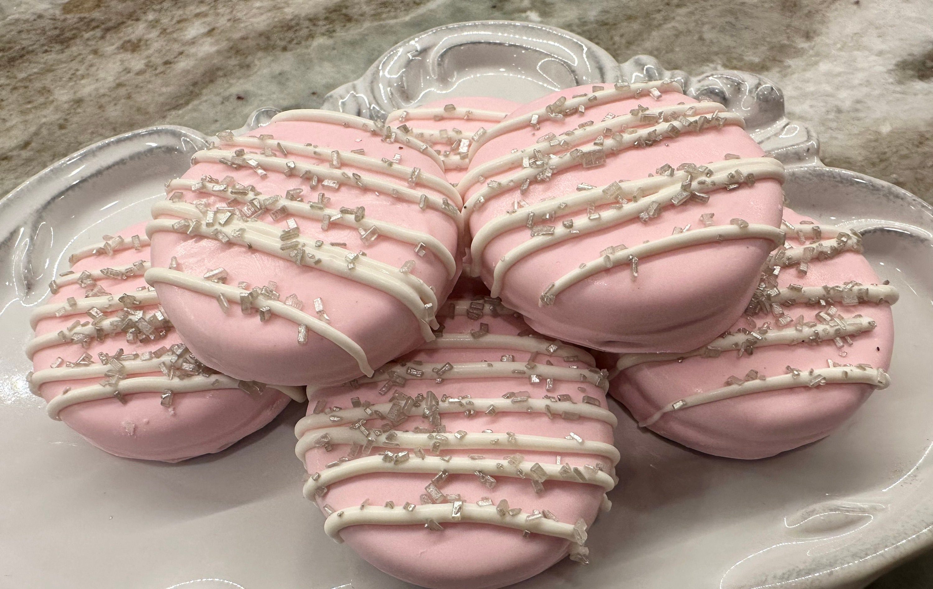 Pastel pink chocolate-covered Oreos with white icing and sprinkles on an ornate white plate, available as a 12-pack for weddings or baby showers.