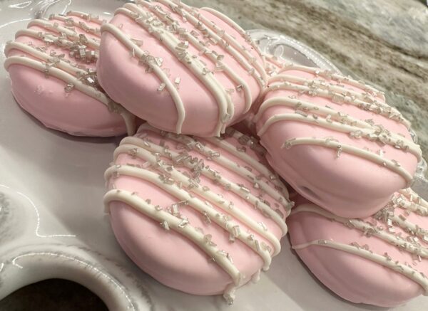 A plate filled with 12 pastel pink chocolate-covered Oreos, topped with white drizzle and silver sprinkles, perfect for wedding or baby showers.