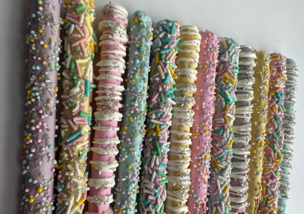 Spring Decorated Chocolate Covered Pretzel Rods, adorned with colorful sprinkles, arranged side by side on a white surface.