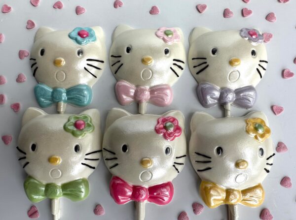 Six lollipops from the "12-Say Hello To My Sparkling Kitty Kat White Chocolate Lollipop Favors" feature cat faces with unique colored bows and flowers, arranged in two rows with small pink heart sprinkles scattered around.