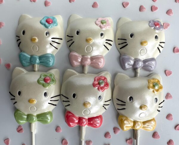 Six "Say Hello To My Sparkling Kitty Kat" white chocolate lollipops decorated with bows and flowers in pastel colors are arranged on a white surface with small pink heart sprinkles.