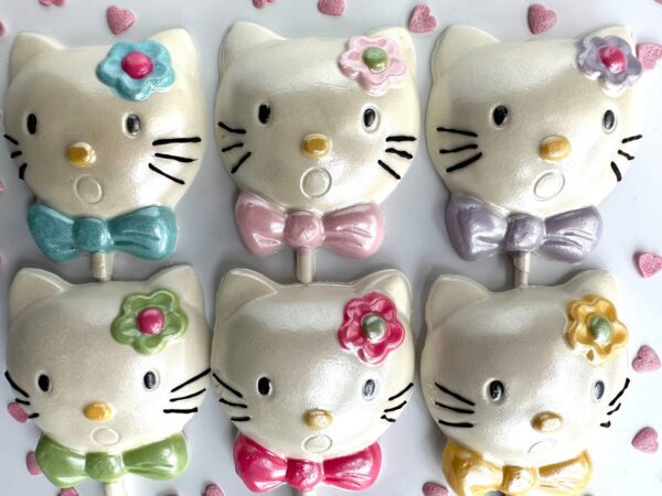 Six cat-shaped white chocolate lollipops with bows and flowers, organized in two rows. Each has a unique colored bow and flower, with small heart-shaped sprinkles scattered around. Product: 12-Say Hello To My Sparkling Kitty Kat White Chocolate Lollipop Favors.