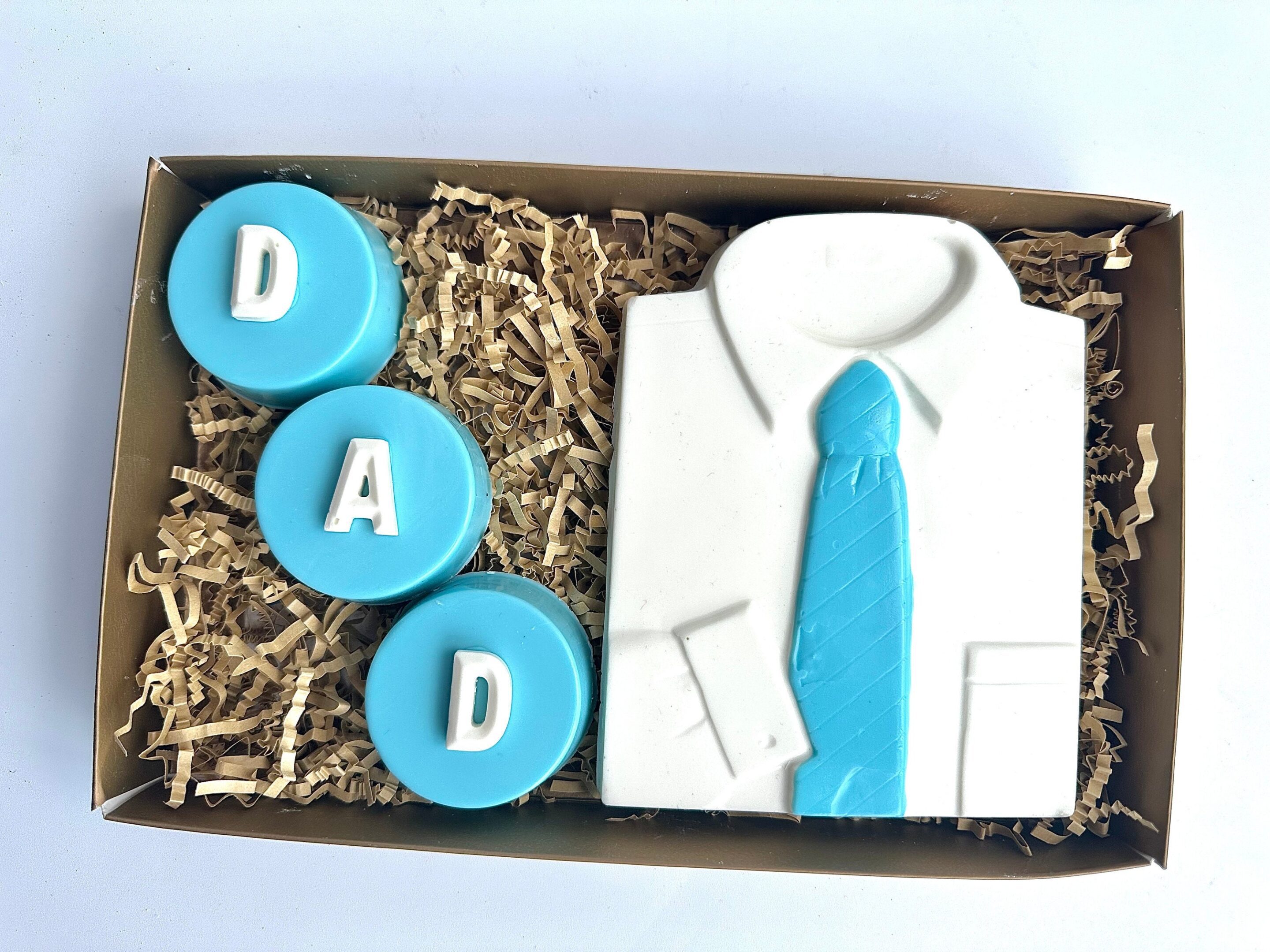 Gift box featuring three blue "DAD" Oreos and a white chocolate shirt with a blue tie, arranged on shredded paper.